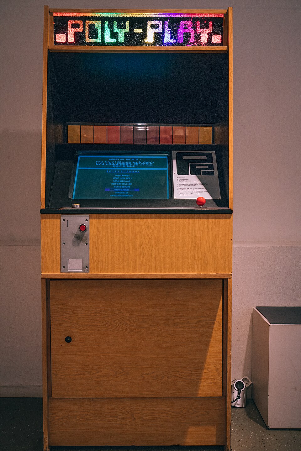 Diese Aufnahme zeigt den Polyplay, den einzigen in der DDR produzierten Arcade-Automaten. Das Gerät im Stehgehäuse ist aus hellem Holzfurnier gefertigt und besitzt eine leuchtende, mehrfarbige "POLY-PLAY"-Anzeige über einem Bildschirm, der aktuell ein blaues Menü mit Spielauswahl anzeigt.