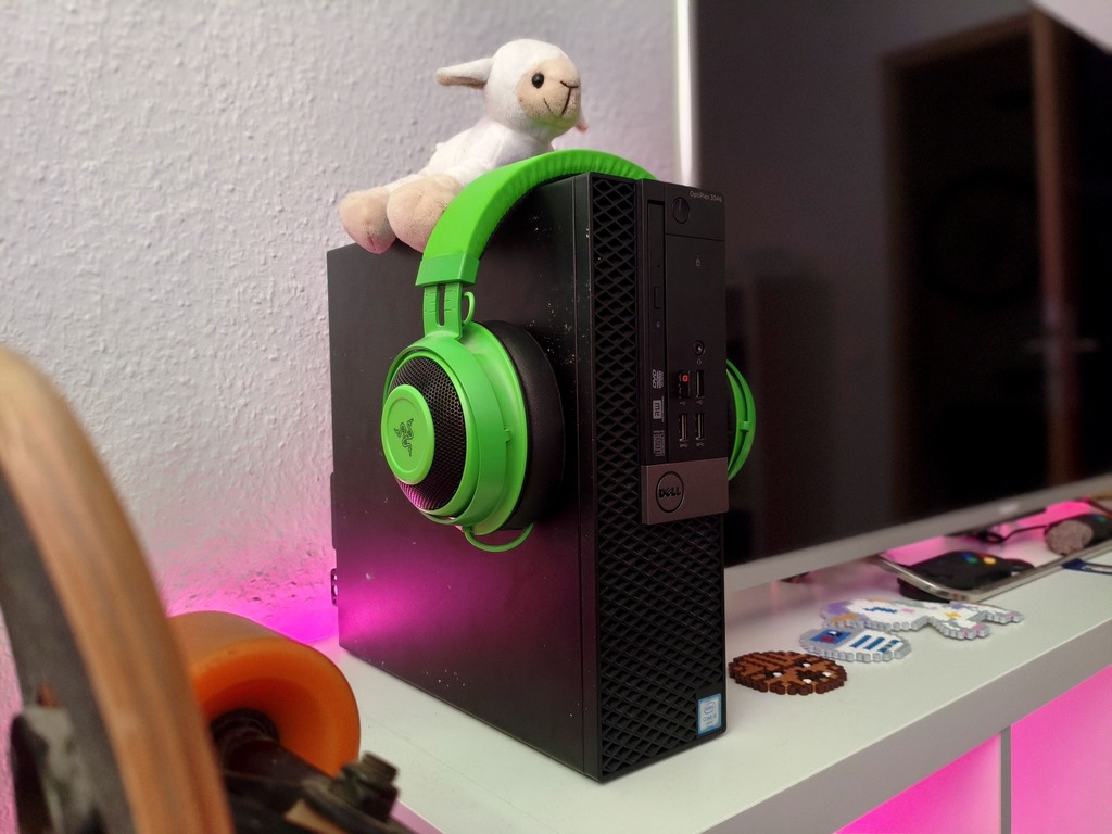 Ein schwarzer Dell OptiPlex Computer steht auf einem weißen Regal. Über die Vorderseite des Gehäuses hängt ein grüner Razer-Kopfhörer, auf der Oberseite des PCs sitzt eine kleine weiße Plüschschaf. Unter und hinter dem Gerät leuchtet eine pinke LED-Beleuchtung. Im linken Vordergrund ist ein Longbard zu erkennen, rechts daneben liegen kleine Pixel-Art-Deko-Figuren aus Perlen.