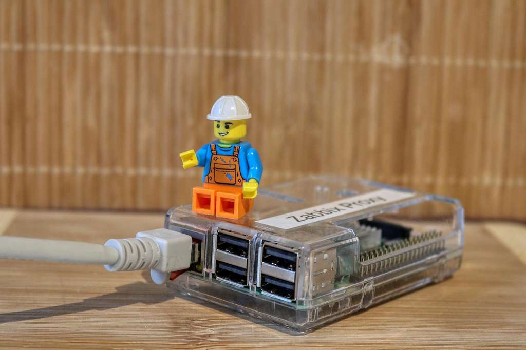 Ein kleines Lego-Männchen mit einem weißen Bauarbeiterhelm, blauer Latzhose und orangefarbenen Schuhen sitzt auf einem durchsichtigen Gehäuse, das einen Raspberry Pi