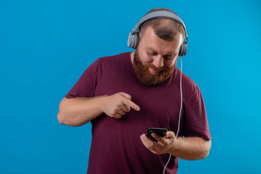Porträt eines bärtigen Mannes mit Kopfhörern, der konzentriert auf sein Smartphone schaut und mit dem Finger auf den Bildschirm zeigt. Der Hintergrund ist ein leuchtendes Blau.