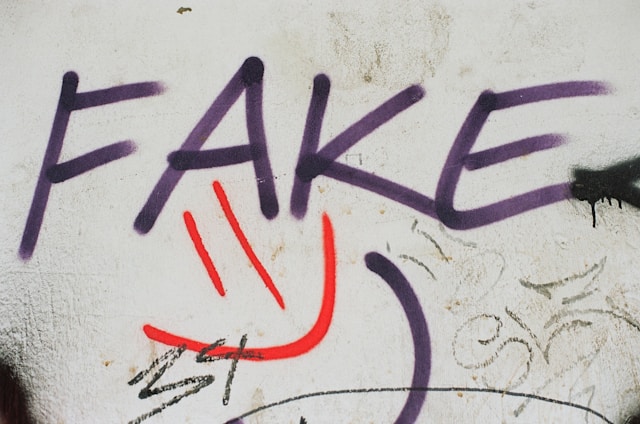 Auf einer abgenutzten, leicht verschmutzten Betonwand ist in großem, lilafarbenem Graffiti das Wort „FAKE“ gesprayt. Die Buchstaben sind grob, aber deutlich lesbar und leicht verwischt, was dem Schriftzug einen urbanen, rebellischen Charakter verleiht. Darunter ist in roter Farbe ein stilisiertes Smiley-Gesicht aufgesprüht – zwei senkrechte Striche als Augen und ein gebogener Strich als lächelnder Mund. Weitere Kritzeleien und Tags in Schwarz und Grau umgeben das Hauptmotiv, was dem Bild einen typischen Street-Art-Flair verleiht.