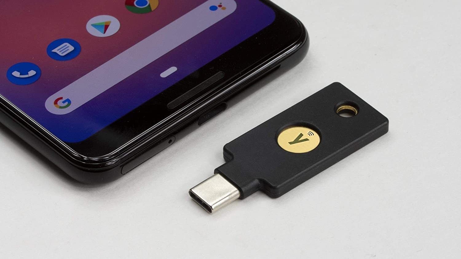 Smartphone und direkt daneben einen YubiKey Sicherheitsschlüssel mit USB-C Anschluss. Der YubiKey ist schwarz, rechteckig und hat in der Mitte ein rundes, goldenes Kontaktfeld mit einem eingravierten „Y“-Logo (dem Yubico-Logo). Über dem Logo sieht man ein kleines NFC-Symbol, das anzeigt, dass der YubiKey auch drahtlos (per NFC) genutzt werden kann.