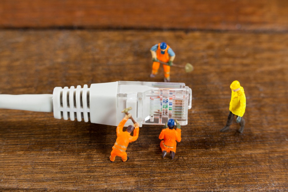 Miniaturarbeiter „reparieren“ symbolisch einen Netzwerkkabelstecker – eine humorvolle Darstellung digitaler Infrastrukturpflege im Kleinformat. Die Szene spielt auf einer Holzoberfläche und kombiniert Technik mit Modellbau-Charme.